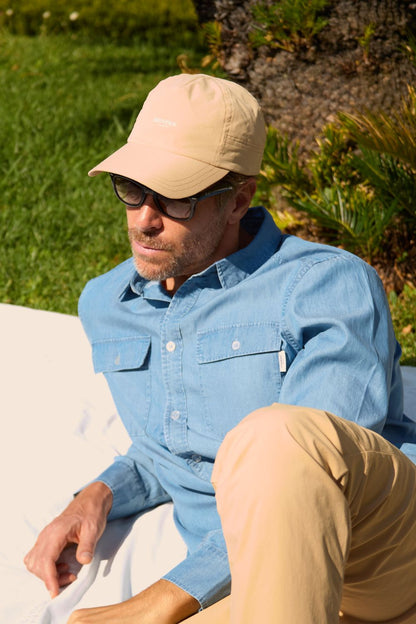 Homme portant une casquette anti-UV beige, lunettes de soleil, assis sur l'herbe en plein air.