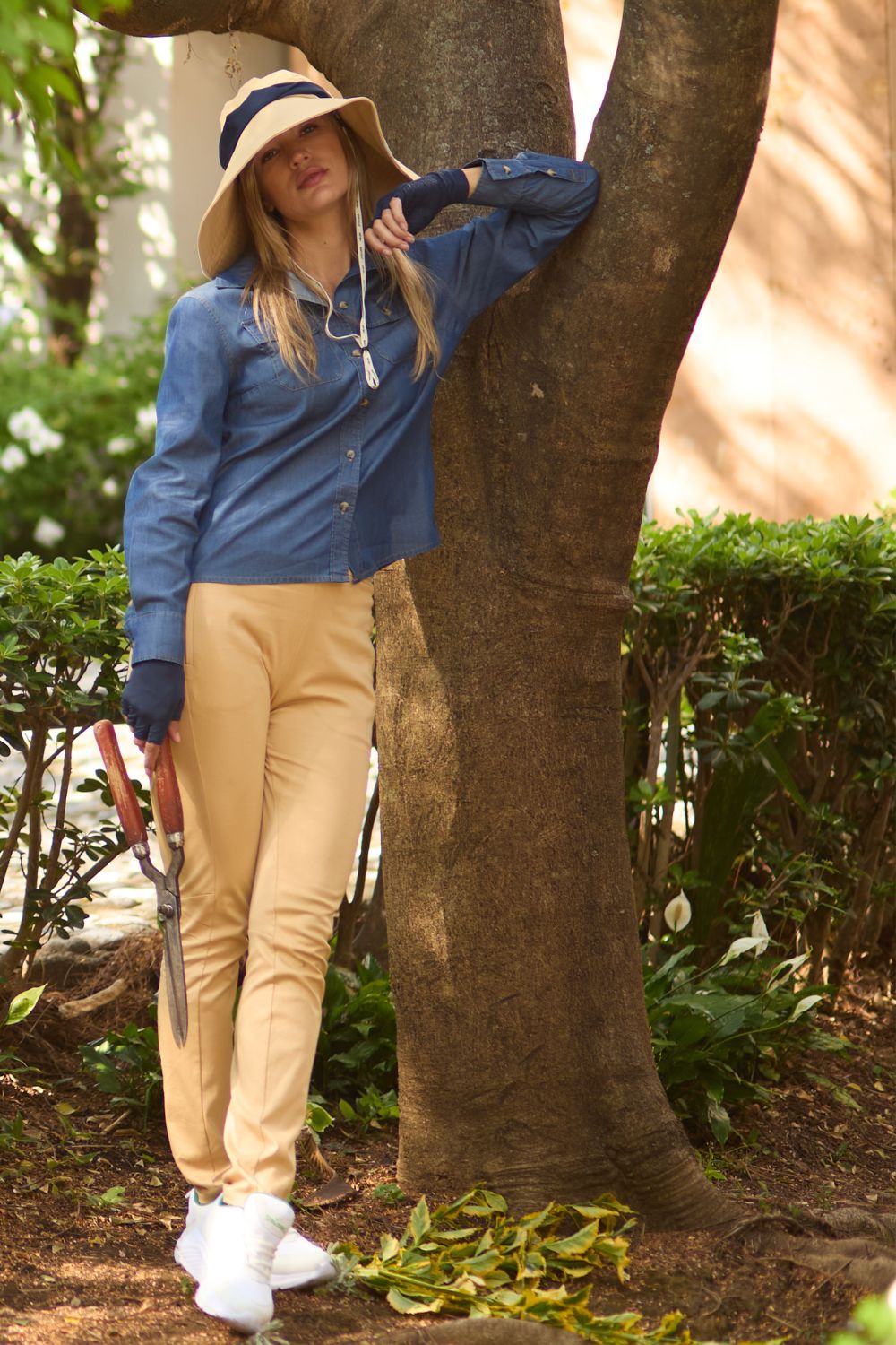 Femme en pantalon anti-UV beige, chemise bleue, chapeau, au jardin, avec protection solaire UPF 50+.